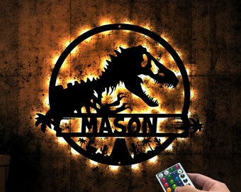 May include: A black metal dinosaur silhouette with a light-up halo and the name "MASON" in the center. The dinosaur is surrounded by palm trees and a mountain. The light is controlled by a remote with multiple color options.