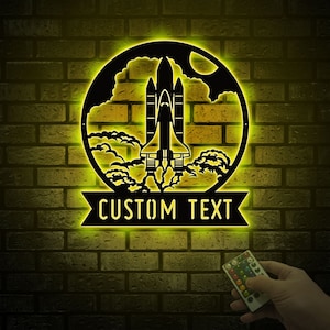 May include: A black metal wall decor with a space shuttle design inside a circle. The shuttle is flying through clouds and the text "CUSTOM TEXT" is displayed below the circle.