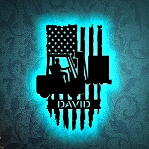May include: A black silhouette of a forklift with a driver inside, superimposed on an American flag with the name "DAVID" below. The image is illuminated with a blue light.