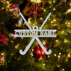 May include: Silver glitter Christmas ornament featuring two crossed hockey sticks with a banner that reads "CUSTOM NAME".