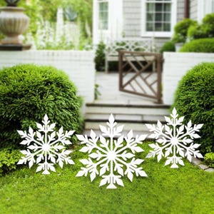 May include: Three white metal snowflake yard decorations in a grassy lawn. The snowflakes are different sizes and have a delicate, intricate design.