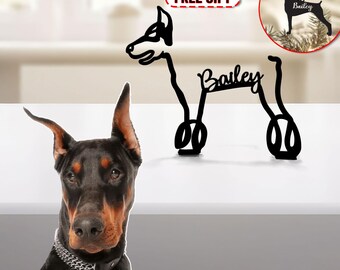 May include: Black metal silhouette of a dog with the name "Bailey" written across its back. The silhouette is a Christmas ornament and is shown on a white background. The text "FREE GIFT" is in a red box above the ornament. A real dog is shown in the lower left corner of the image.