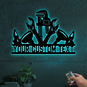 May include: A black metal wall decor with a silhouette of various tools, including a wrench, pliers, and a pipe wrench. The decor is illuminated with blue light and has a customizable text banner that reads "YOUR CUSTOM TEXT".