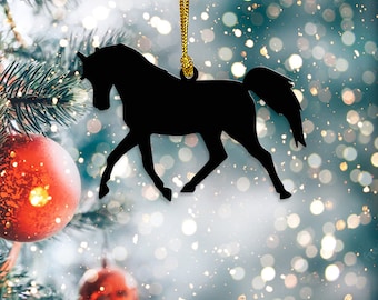May include: Black horse silhouette Christmas ornament hanging from a gold string. The ornament is in the shape of a horse in motion. The background is a blurred image of a Christmas tree with red ornaments and bokeh lights.