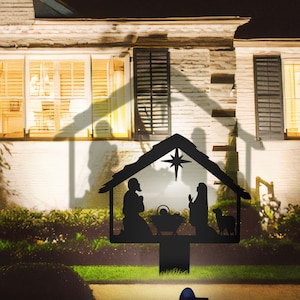 May include: Black metal silhouette of the nativity scene with Mary, Joseph, and the baby Jesus in a manger. The silhouette is lit up with a spotlight, casting a shadow of a house on the background.