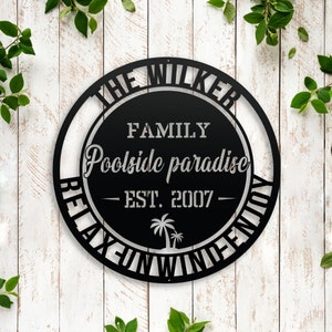 Puede incluir: Se&ntilde;al de metal negro con el texto "The Wilker Family Poolside Paradise - Est. 2007 - Relax, Fun, Wind, Enjoy" en un c&iacute;rculo.