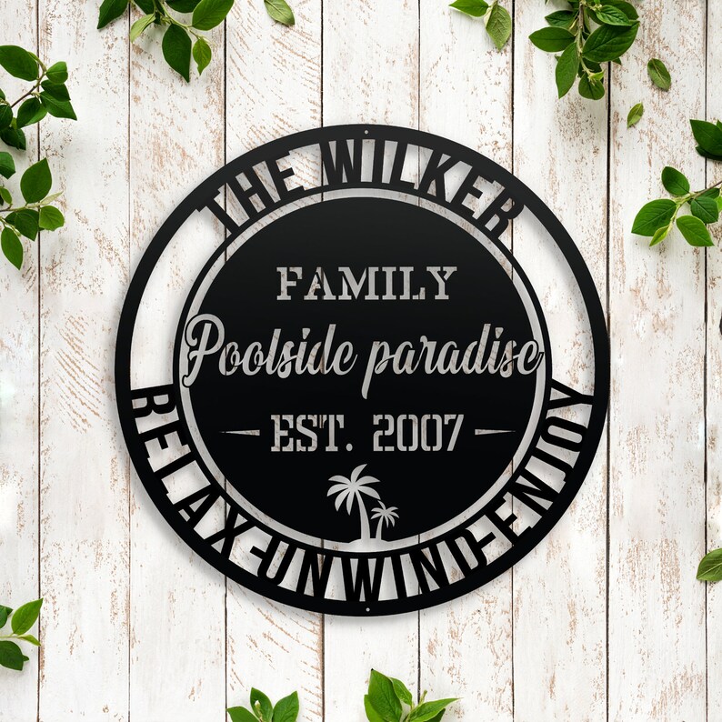 Puede incluir: Se&ntilde;al de metal negro con el texto "The Wilker Family Poolside Paradise - Est. 2007 - Relax, Fun, Wind, Enjoy" en un c&iacute;rculo.