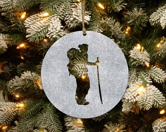 May include: A silver metal Christmas ornament featuring a silhouette of a person hiking with a backpack and a walking stick.