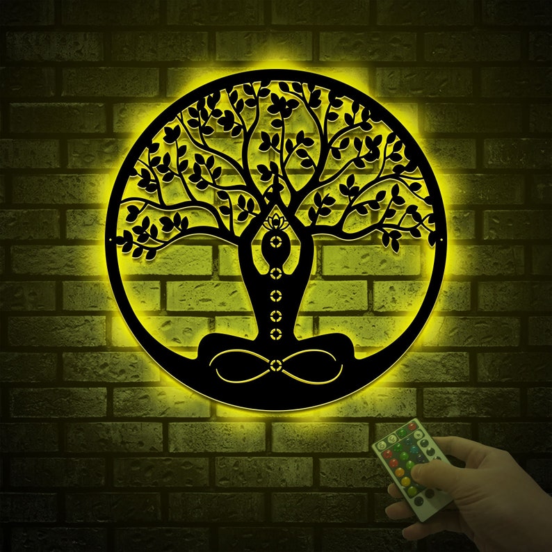 K&ouml;nnte beinhalten: Eine schwarze Wanddekoration aus Metall mit einem Lebensbaum-Design und einer meditierenden Figur in der Mitte. Der Baum hat eine detaillierte Silhouette mit Bl&auml;ttern und &Auml;sten. Die Dekoration wird mit einem warmen gelben Licht beleuchtet.