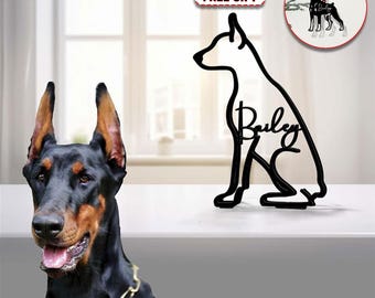 May include: A black and white silhouette of a dog sitting with the name "Bailey" written inside the silhouette. The silhouette is a Christmas ornament. The image also shows a real black and brown Doberman Pinscher dog wearing a collar. The text "FREE GIFT" and "CUSTOM YOUR DOG'S NAME" are also visible in the image.