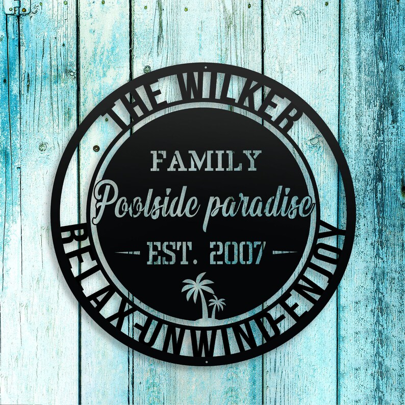 Puede incluir: Un letrero redondo de metal negro con el texto "The Wilker Family Poolside Paradise - Est. 2007 - Relax, Unwind, Enjoy" en fuente blanca. El letrero presenta una silueta de palmera.