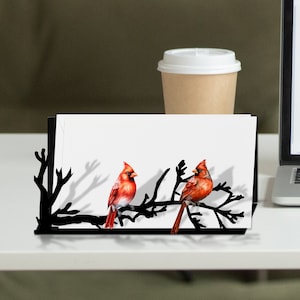 May include: Black metal card holder with a silhouette of a branch with two red cardinals perched on it. The holder is on a white surface with a white coffee cup.