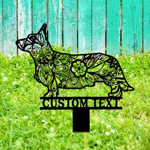 May include: Black metal silhouette of a dog with a floral pattern, standing on a metal stake with a sign that says "CUSTOM TEXT". The dog is in front of a green grassy lawn with a blue wooden fence in the background.