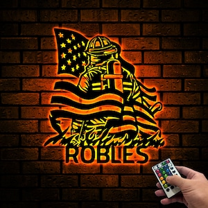 May include: A black silhouette of a firefighter holding an American flag with the name "ROBLES" underneath. The image is lit up in orange and yellow.
