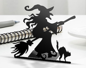 May include: Black metal silhouette of a witch riding a broomstick with a black cat beside her. The witch is wearing a pointed hat and has long flowing hair.