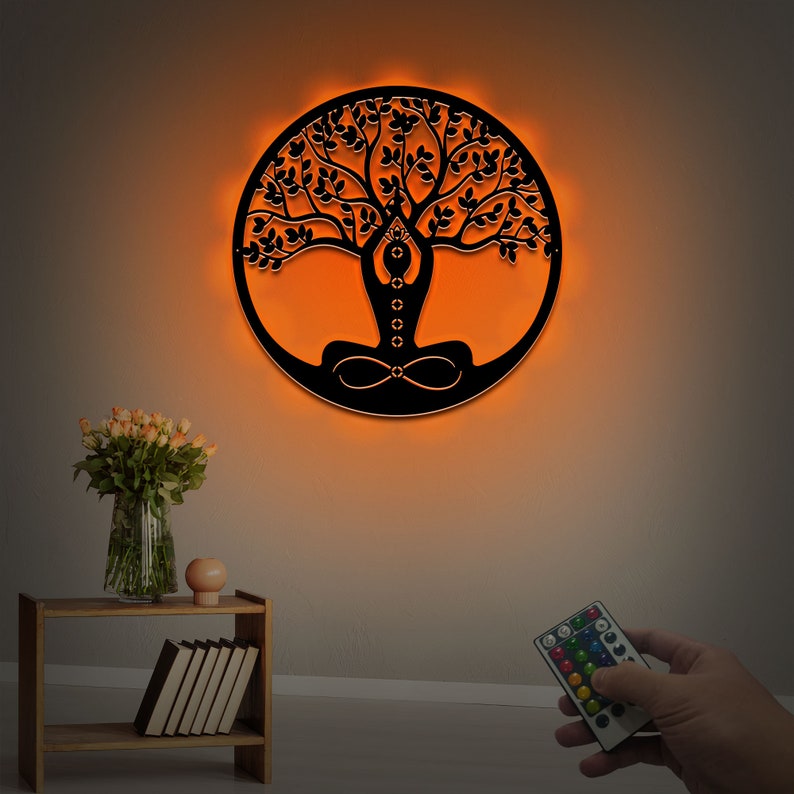 K&ouml;nnte beinhalten: Eine schwarze Wandskulptur aus Metall, die einen Lebensbaum mit einer meditierenden Figur in der Mitte darstellt. Die Skulptur wird mit warmem orangefarbenem Licht beleuchtet und wird mit einer Fernbedienung geliefert.