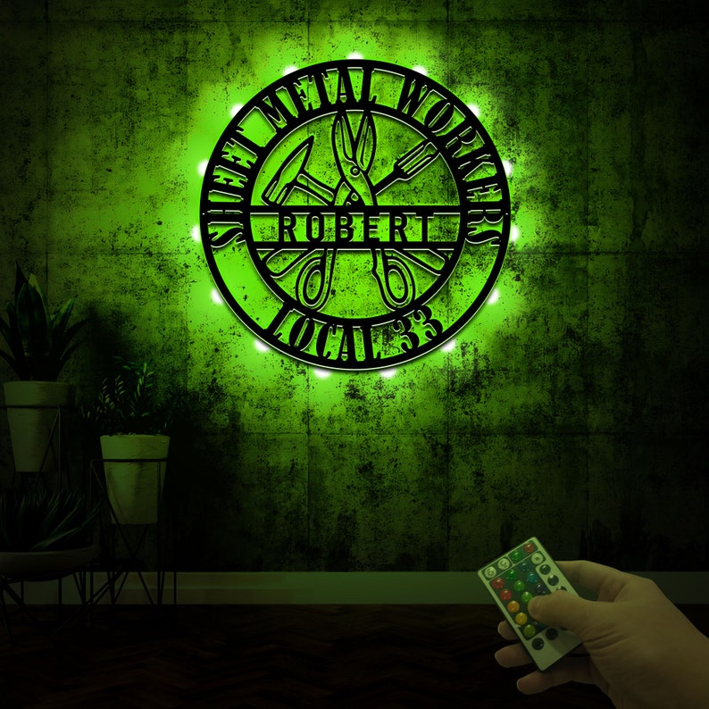 May include: A black metal sign with the words "Sheet Metal Workers Local 33" and the name "Robert" in the center. The sign is illuminated with white lights and is mounted on a green wall.