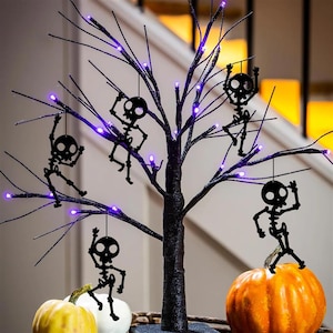 May include: A black Halloween tree with purple LED lights and hanging skeleton decorations. The tree is decorated with small, black skeleton cutouts and sits next to orange and white pumpkins, creating a spooky seasonal display.