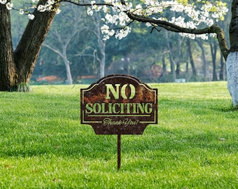 May include: A rusty brown metal sign with the text "NO SOLICITING - Thank You!" in green lettering. The sign is on a green grassy lawn with a white flowering tree in the background.
