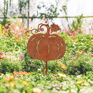 May include: A rusty metal pumpkin silhouette garden stake. The stake is placed in a bed of green plants with pink and yellow flowers.