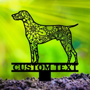 May include: Black metal dog silhouette garden stake with intricate floral and geometric patterns. The dog is standing, and the stake has the words "CUSTOM TEXT" across the bottom. The background is a blurred green.