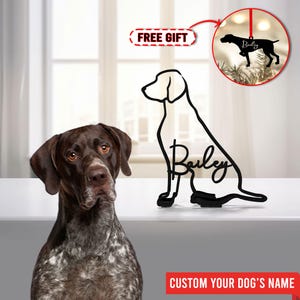 May include: A black metal silhouette of a dog sitting with the name "Bailey" written in cursive. The silhouette is on a white surface with a real dog in the foreground. The text "CUSTOM YOUR DOG'S NAME" is at the bottom of the image.