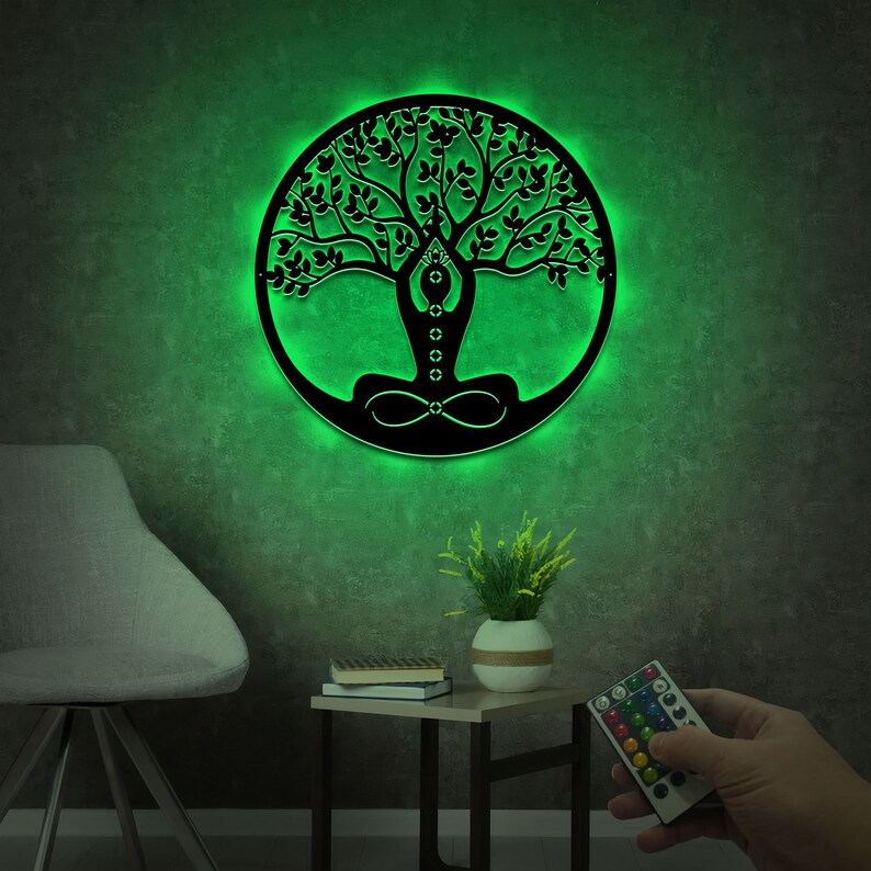 K&ouml;nnte beinhalten: Eine schwarze Metall-Wanddekoration in Form eines Lebensbaums mit einem gr&uuml;nen Licht, das von hinten strahlt. Der Baum hat eine Person, die im Zentrum des Baumes meditiert. Die Wanddekoration h&auml;ngt an einer gr&uuml;nen Wand.