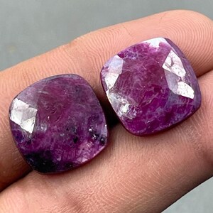 May include: Two cushion-cut gemstones with a deep magenta colour. The gems have a polished surface with multiple reflective facets, creating a sparkling effect. The stones are held in a hand, showcasing their colour and clarity.