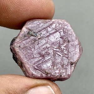 May include: A deep pink ruby crystal with a faceted, hexagonal shape. The crystal has a rough, natural texture and is translucent.