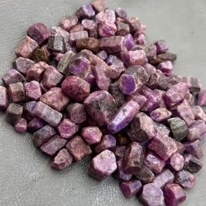 Op de afbeelding: Een stapel ruwe robijn edelstenen in verschillende vormen en maten. De stenen vertonen een reeks kleuren van diep rood tot paarsachtig roze, waarbij sommige donkerdere, bijna bruine tinten vertonen. De ruwe, ongepolijste textuur van de edelstenen is duidelijk zichtbaar.