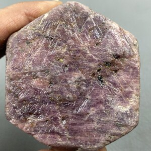 以下が含まれることがあります： 深いピンクがかった紫色の六角形のルビー原石。表面は、目に見える結晶構造と小さな黒いインクルージョンがあり、テクスチャ加工された外観をしています。原石は手で持たれており、その自然な未研磨の状態を示しています。