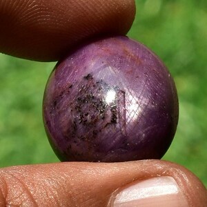 May include: A polished, spherical gemstone held between fingers. The gem is a deep purplish-red color with dark inclusions and a reflective surface. The background is blurred green.