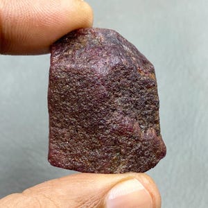 Può includere: Una gemma di rubino grezza e non tagliata, di colore rosso-viola intenso. La pietra naturale ha una superficie ruvida e una forma irregolare. Il rubino è tenuto tra le dita su uno sfondo neutro, non lucido.