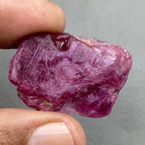 May include: A rough, uncut ruby gemstone held between fingers. The ruby is a deep, saturated pinkish-red color with visible crystalline structures and a slightly reflective surface. The stone has a natural, irregular shape.