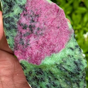 May include: A natural ruby-in-zoisite stone, featuring a central area of bright pink ruby set within a green zoisite matrix. The stone has an organic shape, with black inclusions scattered across the green.