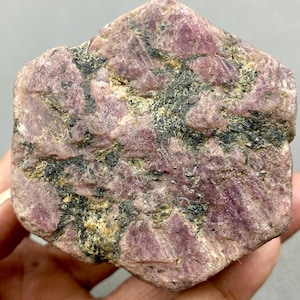 May include: A rough, pink and black ruby gemstone with a natural, unpolished finish. The ruby has a dark, almost black, matrix with pink and red crystals embedded throughout.