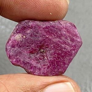 May include: A rough-cut, deep magenta ruby gemstone. The ruby is held between two fingers, showcasing its irregular, hexagonal shape and textured surface. The stone's color is a rich, saturated red-purple.