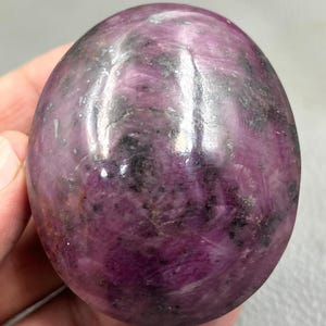 May include: A polished, egg-shaped ruby zoisite stone featuring a smooth, glossy surface with deep magenta and purple hues, interspersed with dark black and grey mineral inclusions throughout the crystalline structure of the gemstone.