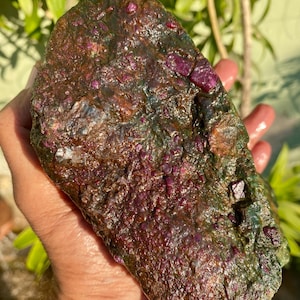 Peut inclure: Une grosse pierre précieuse brute de couleur vert foncé et rouge avec un amas de cristaux de rubis. La pierre précieuse est tenue dans une main.