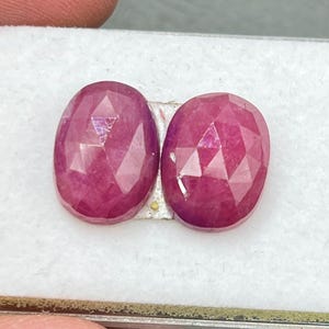 Puede incluir: Dos piedras preciosas ovaladas facetadas de un tono rosa intenso. Las piedras preciosas están colocadas en una caja blanca.