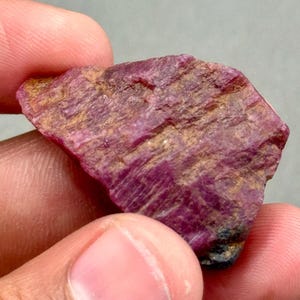 Può includere: Una gemma di rubino grezza, di colore rosso intenso e rosa. La superficie presenta un aspetto strutturato e cristallino con striature più chiare. La gemma è tenuta tra due dita, mostrando la sua forma naturale e non lucidata.