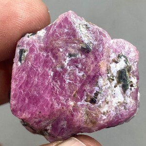 May include: A raw ruby gemstone, displaying a deep pink colour. The surface is textured, with black and white inclusions. The uncut gemstone is held, highlighting its natural, unpolished state.