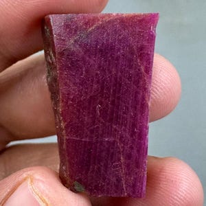 Può includere: Una gemma di rubino grezza, rettangolare, di colore magenta intenso. La pietra ha una superficie ruvida e strutturata con venature interne visibili. Il rubino è tenuto tra le dita, mostrando la sua forma e il suo colore naturali.