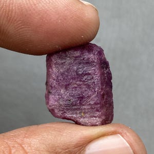 Può includere: Una gemma di rubino grezza, color magenta intenso. Il rubino è tenuto tra due dita, mostrando la sua consistenza naturale non lucidata e il suo colore ricco. La gemma ha una forma rettangolare.