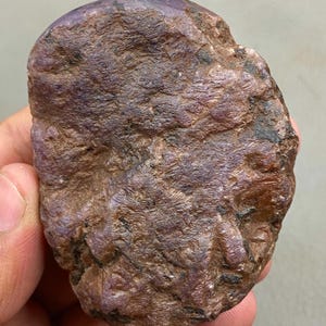 Puede incluir: Una gema de rubí en bruto y sin cortar, de color rojo púrpura intenso. La piedra natural tiene una superficie texturizada con estructuras cristalinas visibles y variaciones de color, sostenida en una mano.