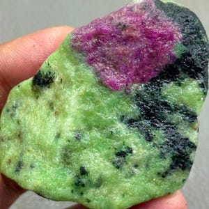 May include: A natural gemstone with a striking fuchsia centre set against a speckled, light green base. Dark black patches are scattered across the stone. The overall texture is rough and natural, giving it a raw, unpolished appearance.