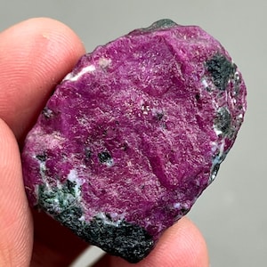 Puede incluir: Una gema de rubí púrpura sin pulir con inclusiones negras y verdes. El rubí es una muestra natural sin pulir.