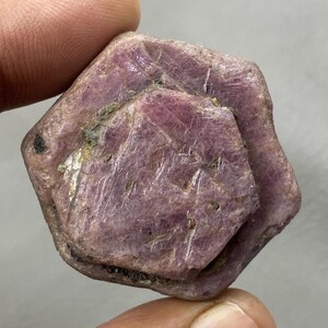 Puede incluir: Una gema de rubí en bruto sin cortar con forma hexagonal. El rubí es de color rojo rosado intenso con inclusiones naturales visibles y una superficie ligeramente irregular. La gema se sostiene entre dos dedos.