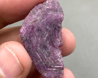 May include: A rough-cut, natural ruby gemstone. The ruby is a deep, vibrant pinkish-purple color with a textured surface. The gemstone is held in a person's hand against a gray background.