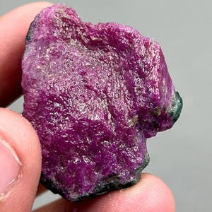Pode incluir: Uma gema de rubi bruta, com uma cor magenta profunda. A superfície tem uma aparência texturizada e cristalina com toques de verde e preto nas bordas. Este espécime mineral natural é segurado em uma mão.